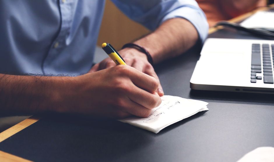 content writing career — Close-up of a man writing notes with a pen next to a laptop on a desk.
