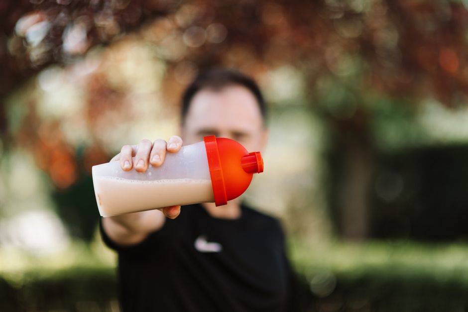 too much protein — Focus on a man holding a plastic protein shake tumbler outdoors with a blurred background.